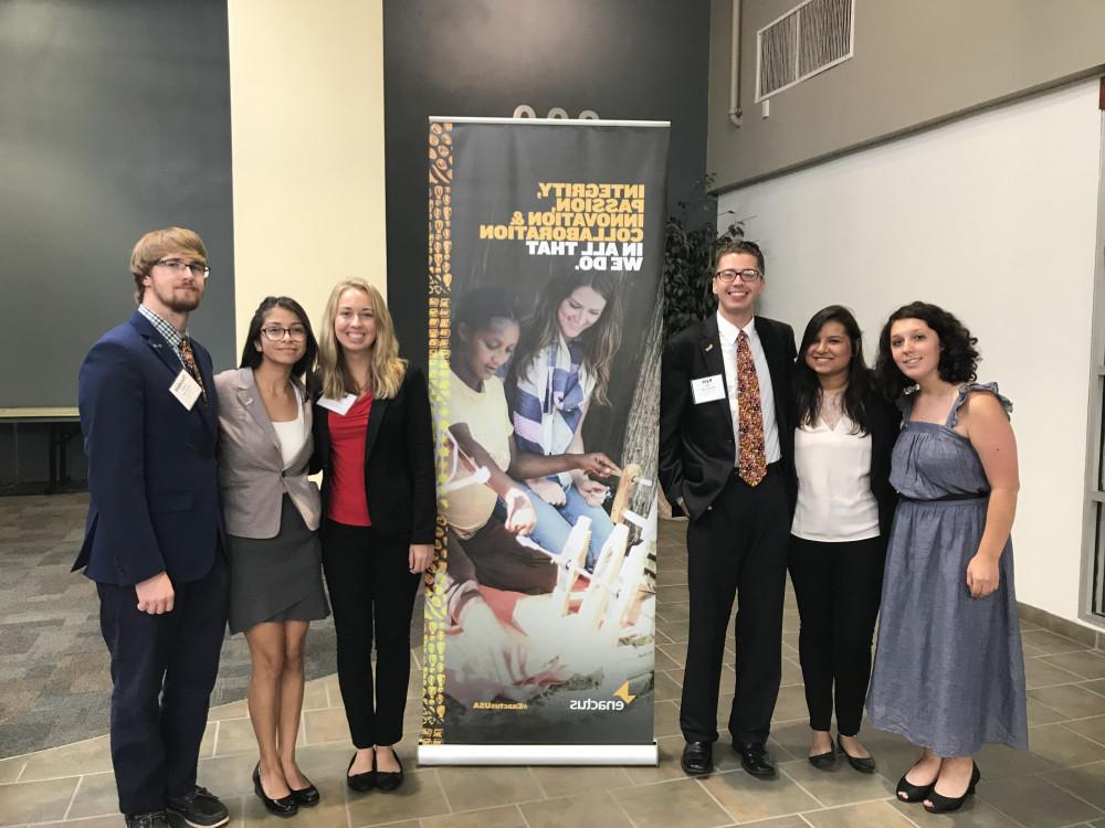 Four female and two male student members of Enactus stand in front of an Enactus banner. 他们穿着正式.